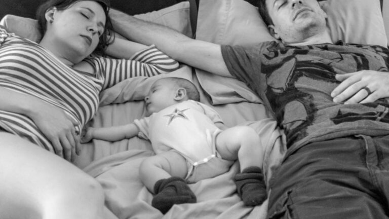 Home 57 A couple lying in bed with their newborn, tender and tired — marriage after baby